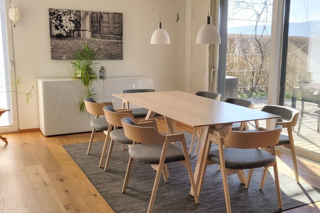 Eine Sitzgruppe bestehend aus Massivholztisch und acht Stühlen. Der Tisch und die Stuhlgestelle sind jeweils aus Eschenholz, weiß geölt. Die Sitzfläche und die Rückenlehne der Sessel sind mit grau meliertem Loden überzogen.