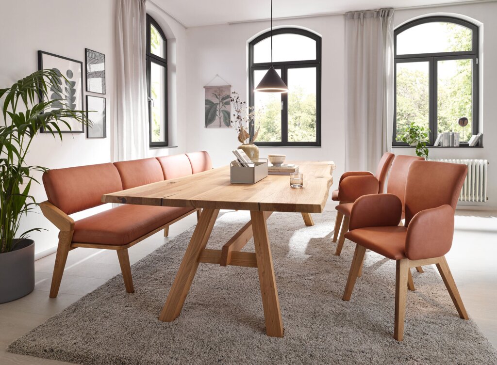 Eine Sitzgruppe mit Tisch aus Eichenholz und Naturkante. Das Tischgestell ist ebenfalls aus Eiche massiv. Eine Sitzbank und drei Stühle mit ebenfalls Massivholz-Gestell und einem terracottafarbenem Lederbezug.