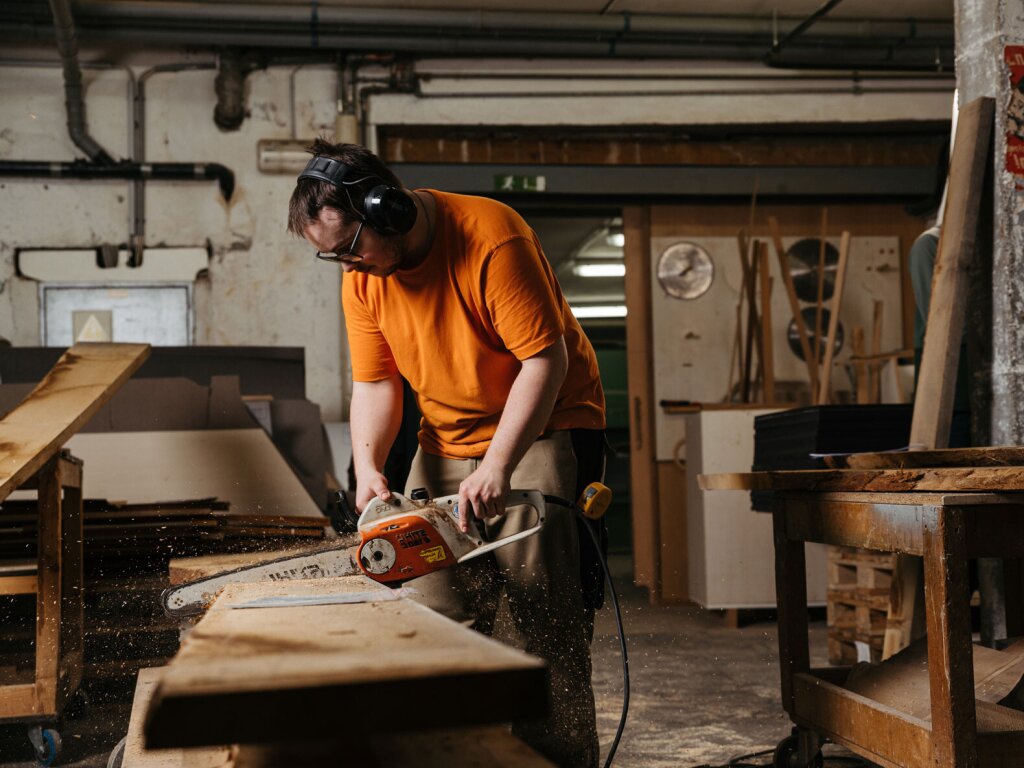 Ein Tischler der Lidauer Tischlerei schneidet mit der Motorsäge massive Zirbenholz - Pfosten zu. Er trägt Gehörschutz, Holzstaub wirbelt durch die Luft, der Tischler arbeitet konzentriert und mit Hingabe. Die Werkstattumgebung wirkt sehr natürlich und authentisch.
