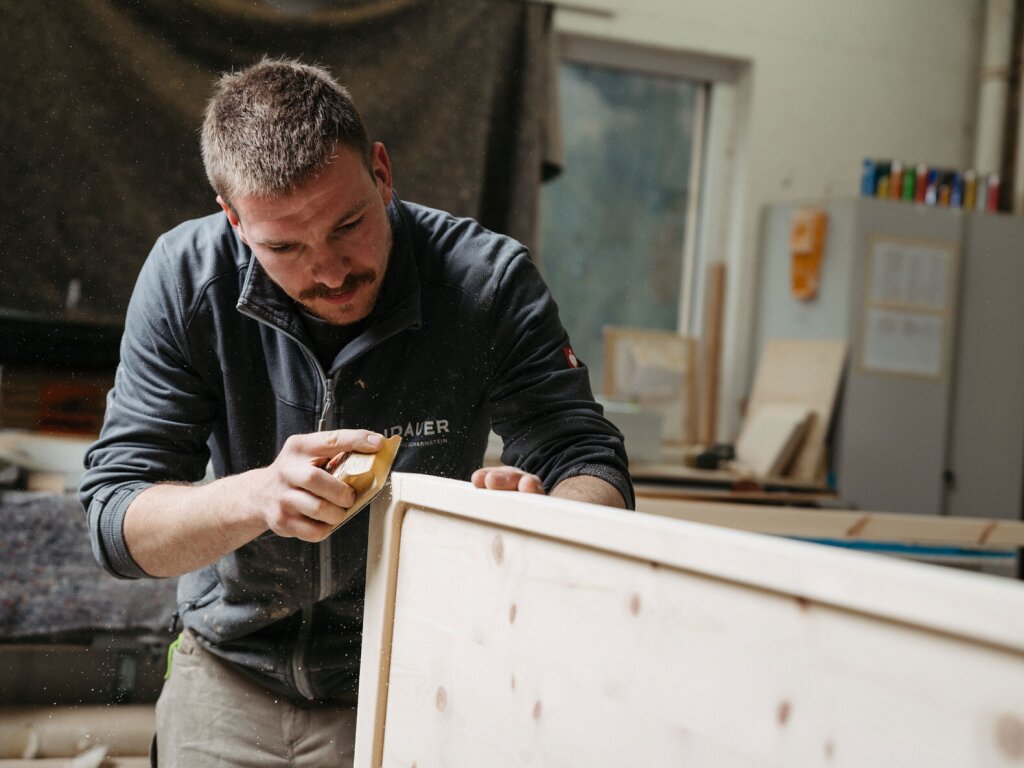 Ein Tischler der Firma Lidauer arbeitet konzentriert an einem Zirbenholzbett. Mit konzentriertem Blick schleift er eine dezente Rundung an der Kante des Betthauptes aus naturbelassener Zirbe. Holzstaub wirbelt in der Luft, die Werkstattatmosphäre hat einen dezenten Retrocharakter.