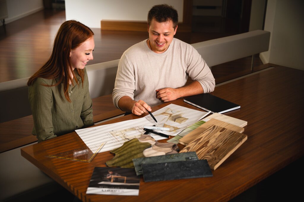 Tischlermeister Sebastian und Planerin Isabel bei der Besprechung eines individuellen Kundenprojekts. Auf dem Tisch liegen Entwurfszeichnungen, Holzproben und Materialmuster in warmen Naturtönen. Die Szene zeigt den kreativen Prozess von der Idee bis zur Planung – persönliche Beratung und individuelle Gestaltung stehen im Mittelpunkt. Eine moderne Arbeitsatmosphäre, die Handwerk, Design und Kundenwünsche verbindet.