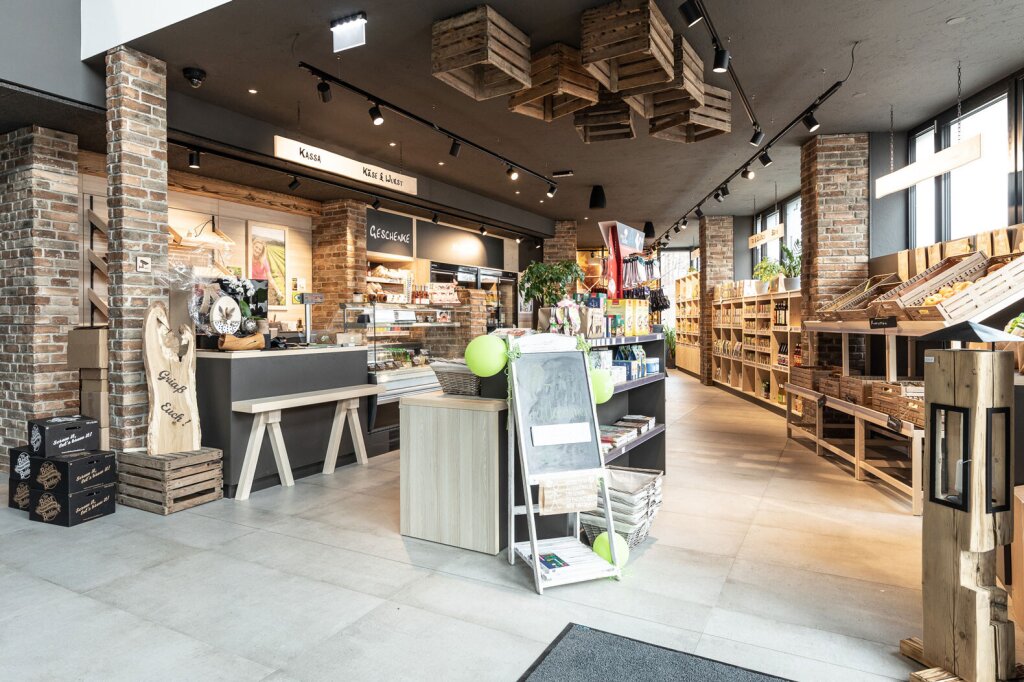 Ein heller Hofladen mit moderner Kassa, Sitzbank und maßgefertigten Holzregalen. In der Mitte ein Regal mit Infotafel und saisonalen Produkten, rechts Obstkisten entlang der Fensterfront. Liebevolle Details wie hängende Holzkisten unter der Decke, dekorative Tafeln und kleine Gestaltungselemente verleihen dem Verkaufsraum eine warme, persönliche Note.