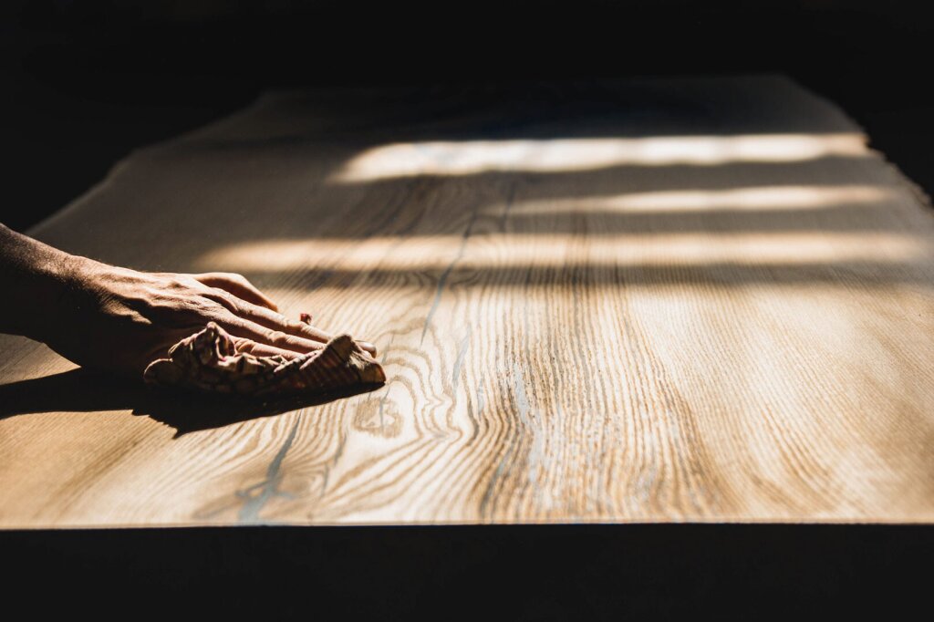 Eine Hand pflegt eine große Massivholzplatte mit einem Tuch im warmen Sonnenlicht. Die Holzmaserung strahlt natürlich und lebendig, während die sanfte Behandlung die Oberfläche schützt und ihre Langlebigkeit unterstreicht. Das Bild vermittelt den Gedanken, dass regelmäßige Pflege nicht nur das Möbel erhält, sondern auch ein nachhaltiger Umgang mit Holz ist.