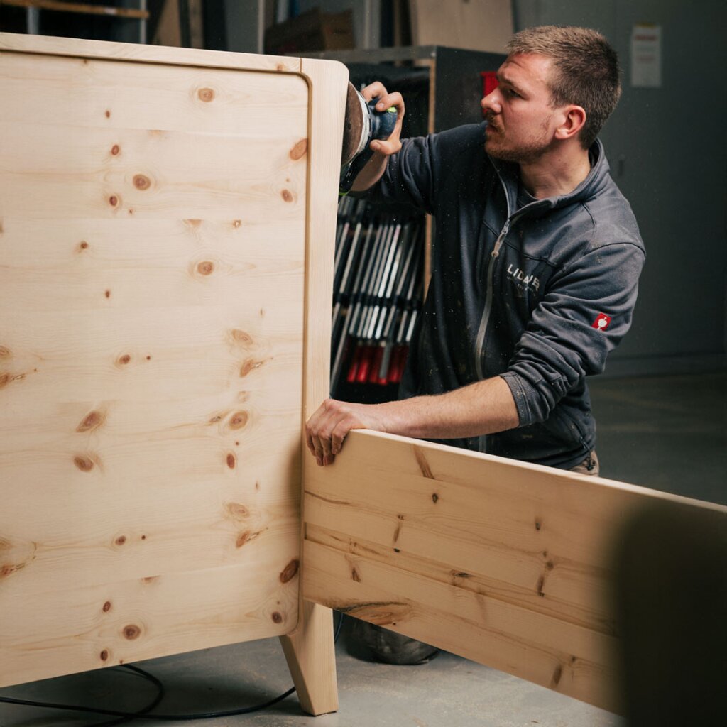 Ein Tischler der Firma Lidauer schleift sorgfältig das Kopfteil eines Bettes aus hellem Zirbenholz. Holzstaub liegt in der Luft, die Szene zeigt den präzisen handwerklichen Prozess, der ein Tischlermöbel einzigartig macht.