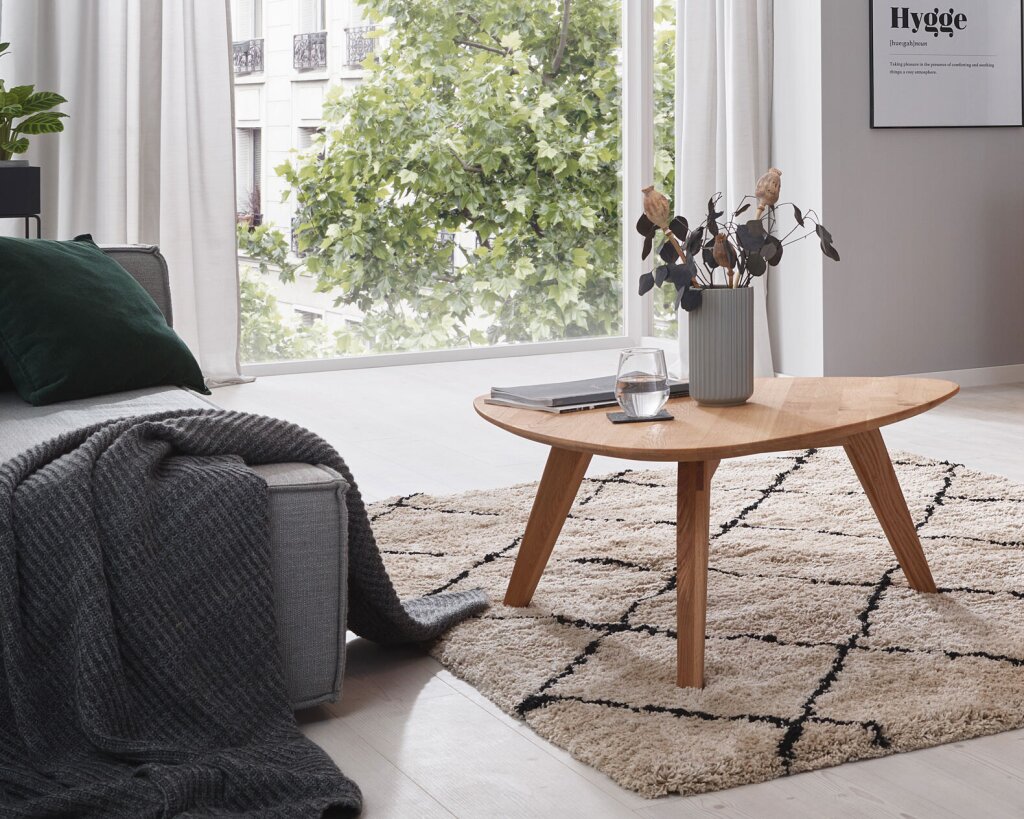 Ein asymmetrischer Couchtisch aus Massivholz in einem modernen Wohnzimmer mit grauer Couch und wohnlichem Teppich. Ein großes Fenster zeigt den Ausblick aus einer Stadtwohnung.