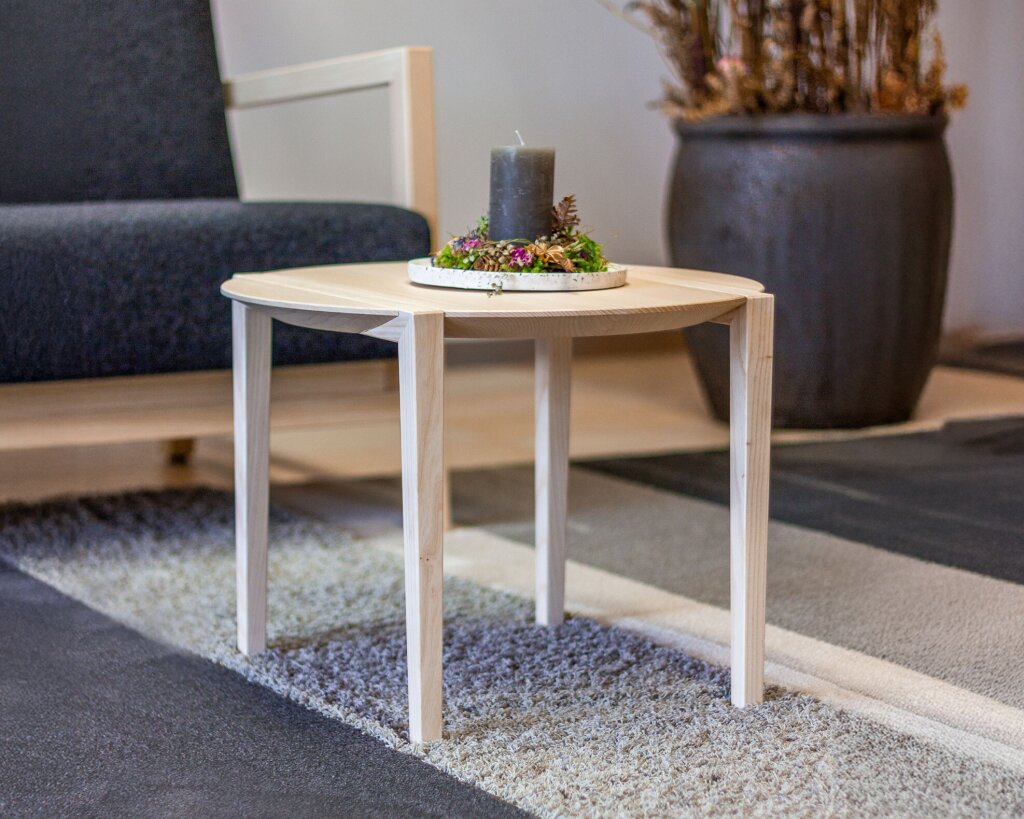 Zierlicher Couchtisch aus hellem Massivholz mit feiner Kante und viereckiger Grundform, in die raffiniert eine runde Tischplatte eingearbeitet ist, platziert auf einem Teppich mit Strukturverlauf. In der Mitte des Tisches steht eine dekorative Schale mit Moos, Zapfen, Blüten und einer dunklen Stumpenkerze. Der Hintergrund zeigt einen gepolsterten Holzsessel und eine große schwarze Keramikvase – ein harmonisches Ensemble aus Natur, Handwerk und Wohnlichkeit.