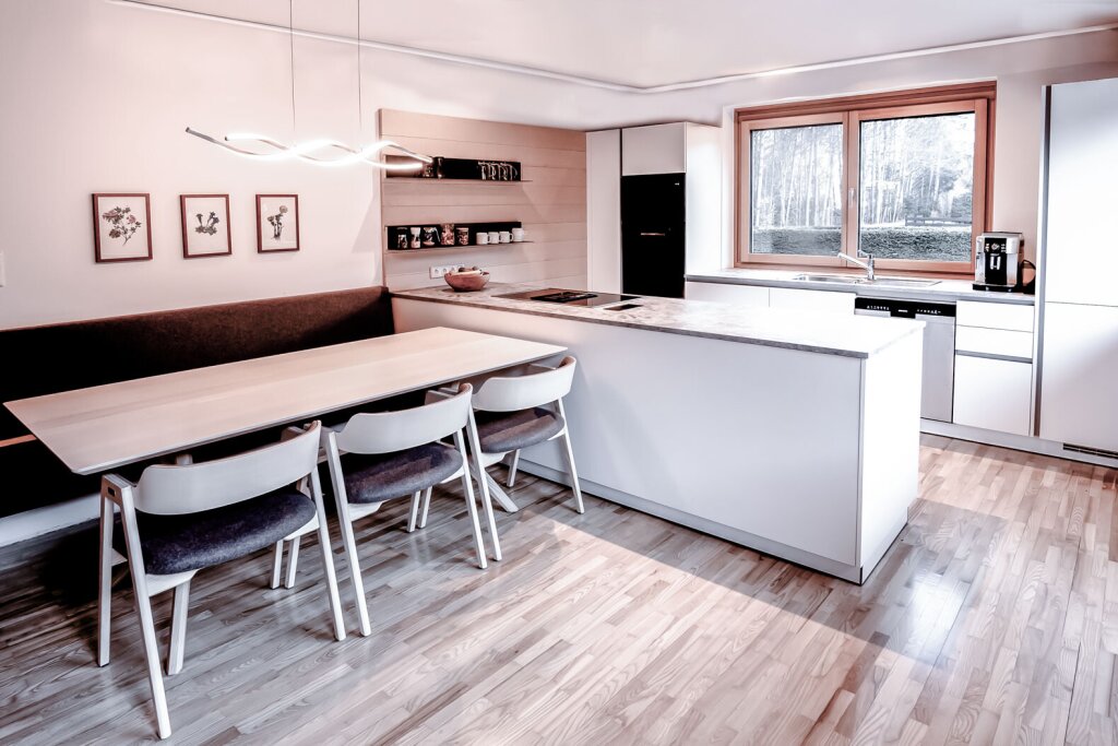 Moderne Wohnküche mit heller Kochinsel, grauer Arbeitsplatte und gemütlicher Sitzecke mit Polsterbank und Stühlen aus Esche mit Lodenpolsterung. Große Fenster bringen viel Tageslicht in den Raum.