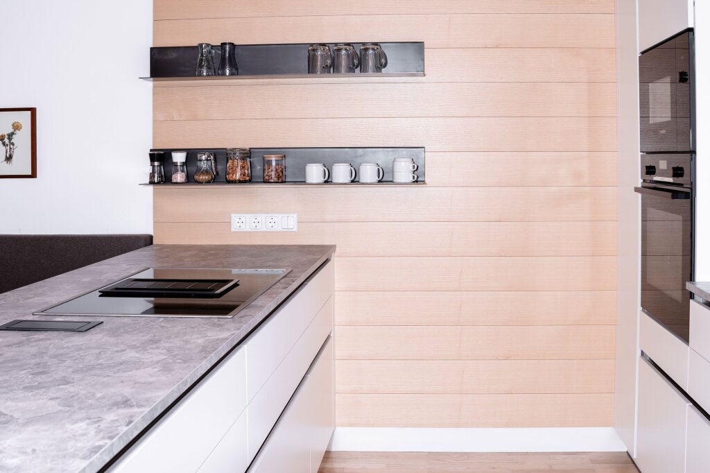 Detailaufnahme moderner Küche mit grauer Stein-Arbeitsplatte und Kochfeld. Offene Regale vor einer Holzrückwand zeigen Gläser, Tassen und Vorratsgläser.