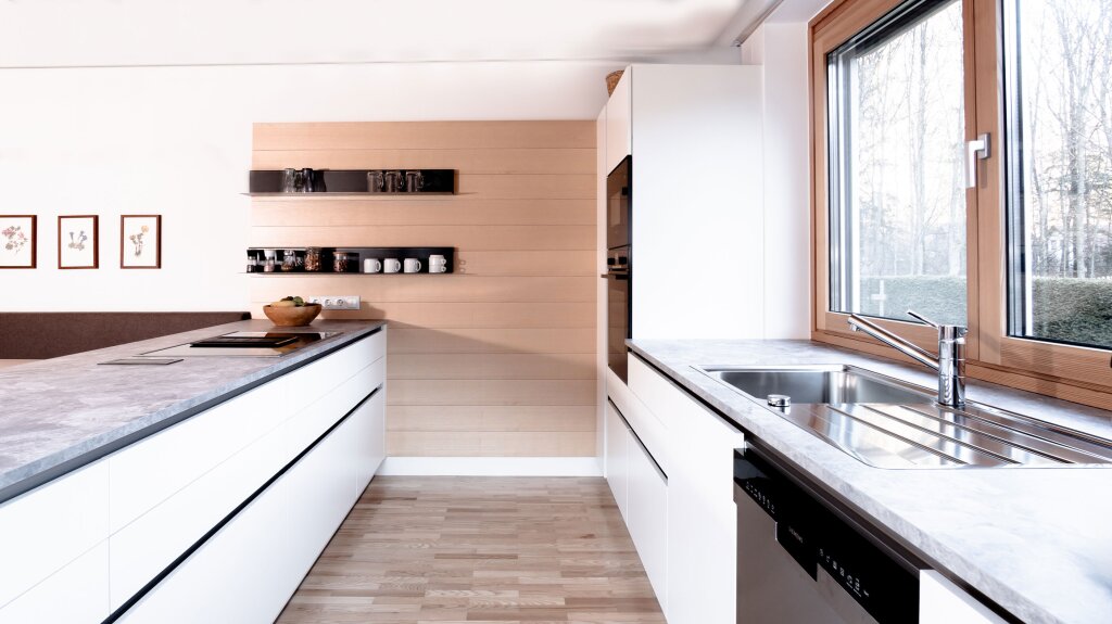Helle Küche mit viel Tageslicht, weißen Fronten und grauen Arbeitsplatten. Holzdetails und offene Regale schaffen eine warme, moderne Wohnatmosphäre.