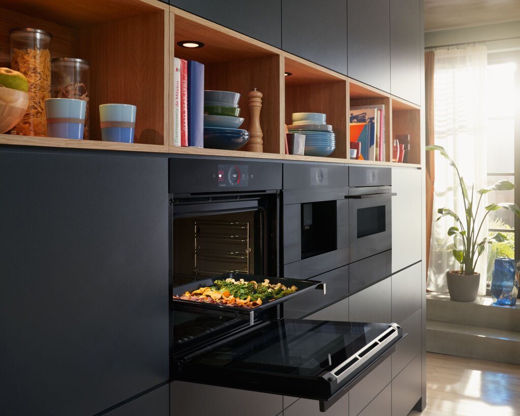 Moderne Einbauküche mit trendigen matten Küchengeräten – Backofen und Dampfgarer in edlem Schwarz-Matt. Offene Holzregale mit Geschirr und Kochutensilien lockern die Fronten auf und schaffen wohnliche Akzente. Tageslicht fällt durch große Fenster und verleiht dem Raum eine einladende, moderne Atmosphäre.