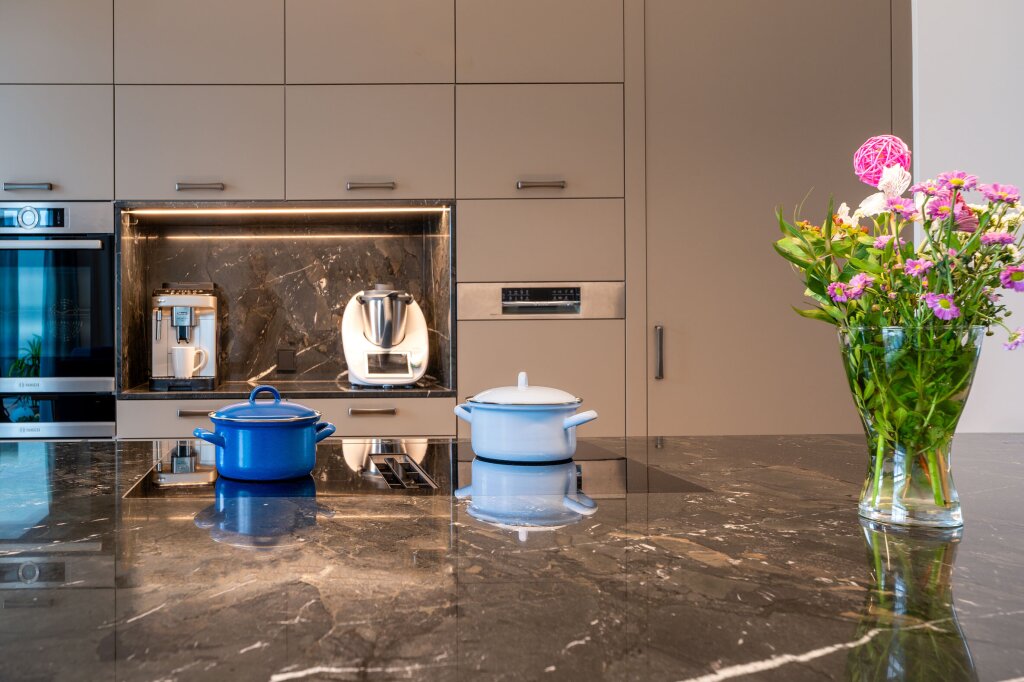 Detailansicht einer modernen Maßküche mit Kücheninsel in dunkler Marmoroptik. Zwei Töpfe stehen auf dem Kochfeld, während im Hintergrund Küchengeräte wie Kaffeemaschine und Thermomix in eine schwarze Nische mit Marmor-Rückwand integriert sind. Mattgraue Fronten, klare Linien und hochwertige Materialien schaffen eine elegante, funktionale Atmosphäre.