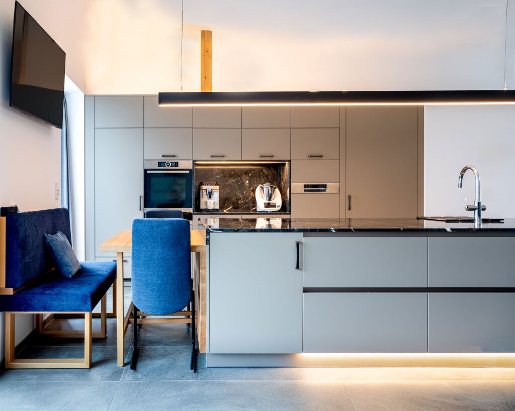 Frontansicht einer modernen Wohnküche nach Maß mit Kücheninsel und integrierter Sitzbank. Mattgraue Fronten, schwarze Stein-Arbeitsplatte und durchgehende LED-Lichtleiste sorgen für Eleganz. Holzdetails, blaue Polsterstühle und hochwertige Einbaugeräte schaffen eine wohnliche, funktionale und repräsentative Atmosphäre.