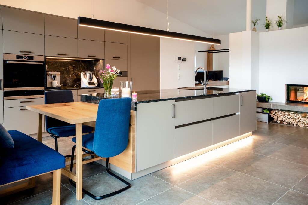 Wohnliche Maßküche mit integrierter Sitzbank und Esstisch – moderne Kücheninsel mit dunkler Stein-Arbeitsplatte und Unterbeleuchtung. Mattgraue Fronten, hochwertige Einbaugeräte und harmonische Holzdetails verbinden Funktionalität mit Eleganz. Blaue Polsterstühle und ein Kamin im Hintergrund schaffen eine warme, einladende Atmosphäre.