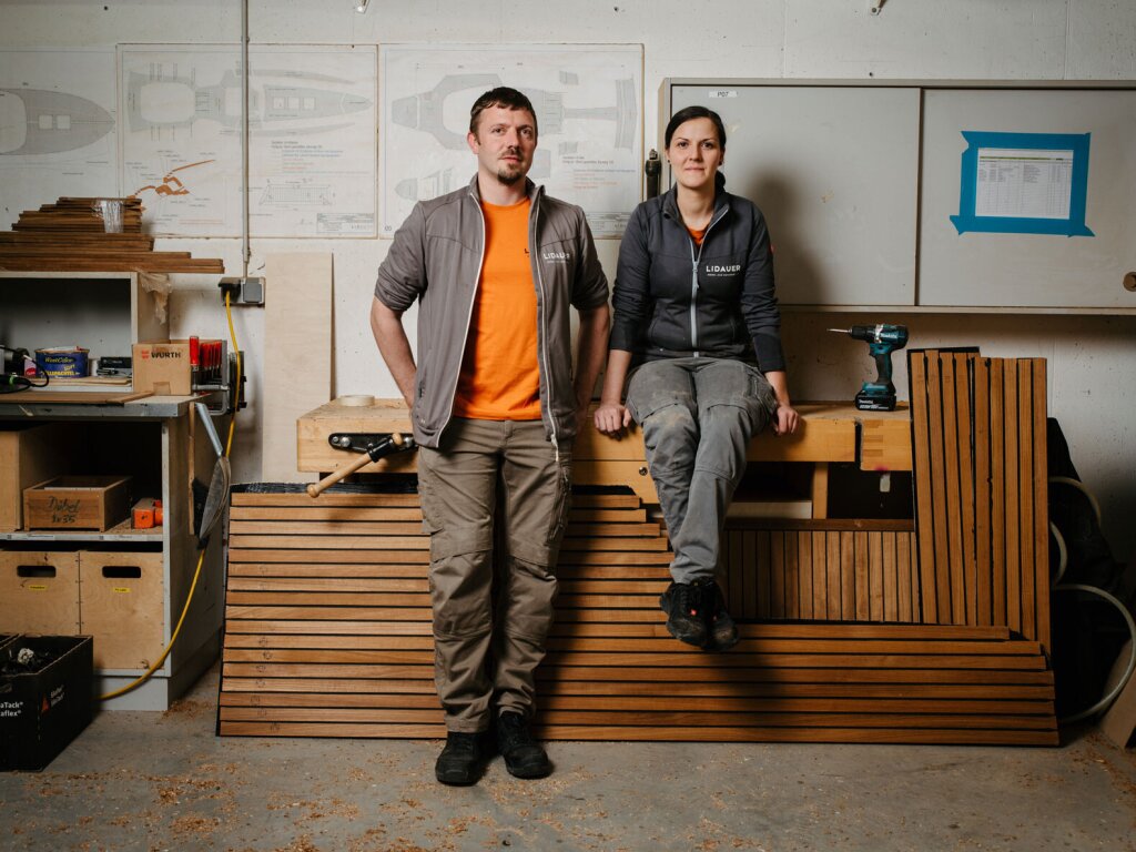 Zwei Tischler in der Bootsbauwerkstätte. Die Tischlerin sitzt auf der Werkbank, der Tischler lehnt neben ihr. Beide blicken direkt freundlich in die Kamera.